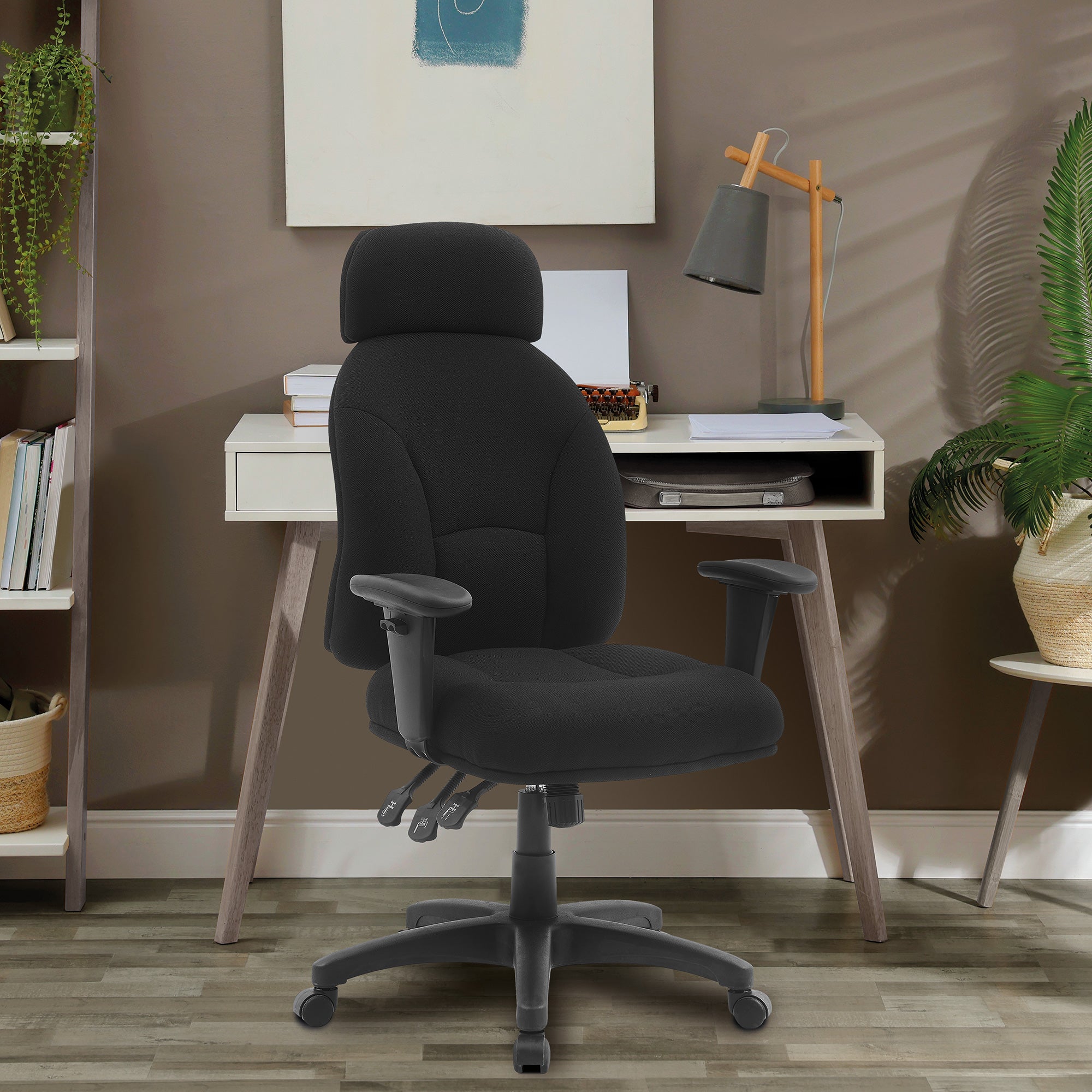 Black office chair in front of a white desk with a lamp and books in a room with a brown wall and plants.