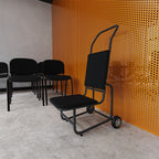 A black chair trolley with a tall curved backrest stands in front of a bright orange perforated metal wall, with additional black chairs lined up against a grey wall on a concrete floor.