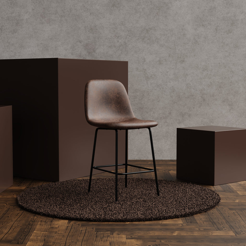 A brown leather bar stool with a black metal frame sits on a round shag rug in a styled interior scene with wood flooring, textured gray walls, and dark brown display blocks.