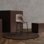 A brown leather bar stool with a black metal frame sits on a round shag rug in a styled interior scene with wood flooring, textured gray walls, and dark brown display blocks.