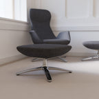 A modern dark grey upholstered ottoman with a chrome star base in the foreground, with matching chairs partially visible in a bright room with white walls and beige carpet.