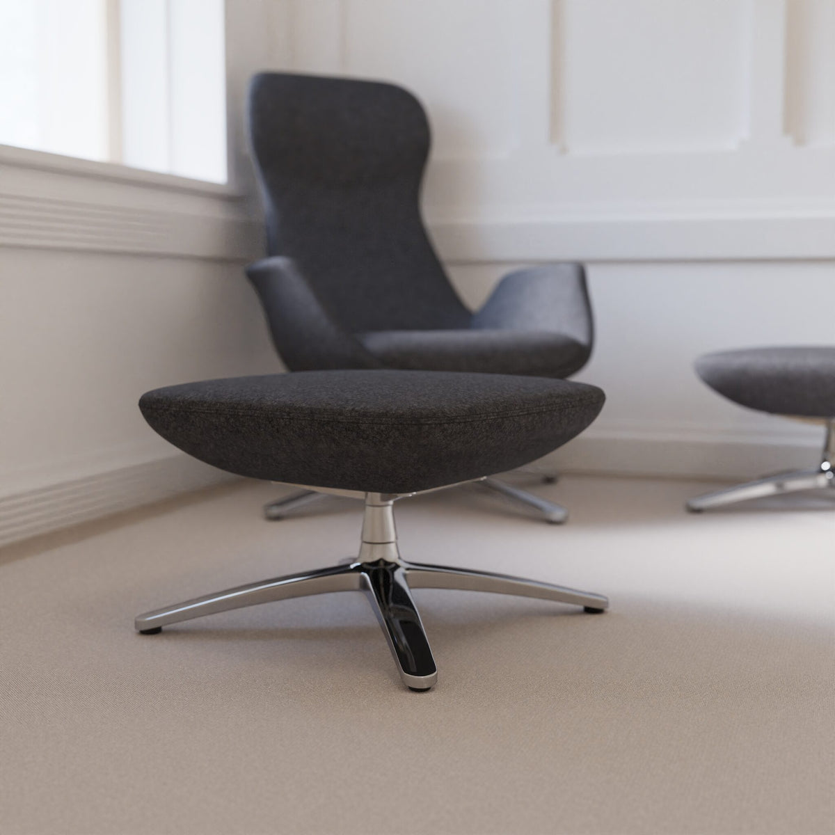 A modern dark grey upholstered ottoman with a chrome star base in the foreground, with matching chairs partially visible in a bright room with white walls and beige carpet.