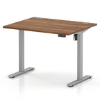 An adjustable standing desk with a medium brown wood-grain desktop and a silver metal frame with dual cable grommets on the top surface.