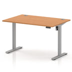 An adjustable height standing desk with a light oak wood grain top and a silver metal frame with T-style legs, shown on a white background.