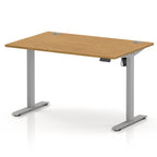 An adjustable height standing desk with a light oak wood grain top and a silver metal frame with T-style legs, shown on a white background.