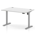 A white height-adjustable standing desk with a silver metal T-style base, featuring cable grommets on the top surface and a control panel on the right side.