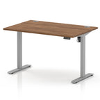An adjustable height standing desk with a medium brown wood-grain desktop and a silver metal frame with T-style legs, shown on a white background.