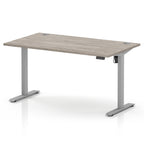 An adjustable height standing desk with a light grey wood-grain desktop and a silver metal frame with T-style legs, shown on a white background.