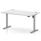 A modern white height-adjustable standing desk with a sleek gray metal T-style frame, featuring a small control panel on the right side and circular cable grommets on the top surface.