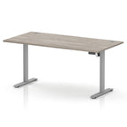 An adjustable height standing desk with a light grey wood-grain desktop and a silver metal frame with T-style legs, shown on a white background.