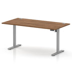 An adjustable height standing desk with a medium brown wood-grain desktop and silver metal legs, shown on a white background with a soft reflection.