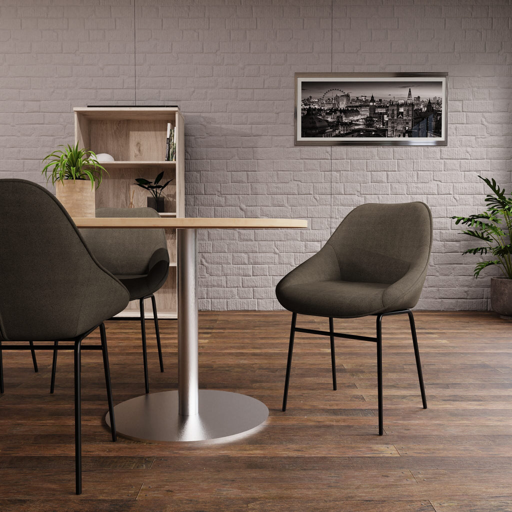 A modern dining setup featuring a round table with a silver pedestal base and three upholstered chairs in a neutral grey-green fabric, set against a light grey brick wall and dark wood flooring.