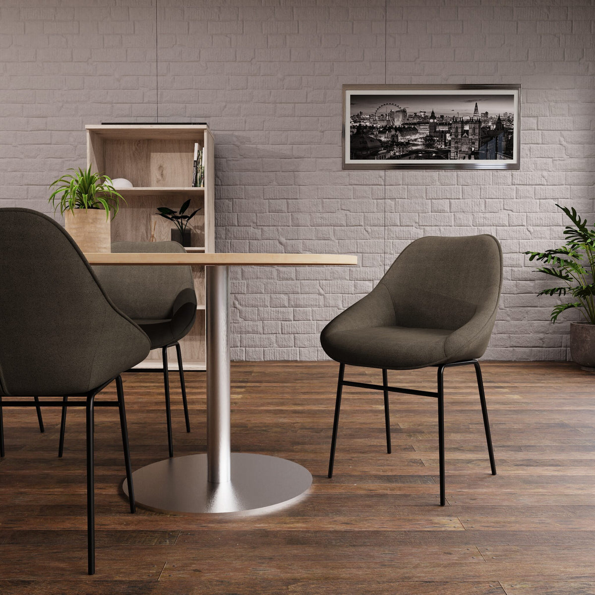 A modern dining setup featuring a round table with a silver pedestal base and three upholstered chairs in a neutral grey-green fabric, set against a light grey brick wall and dark wood flooring.