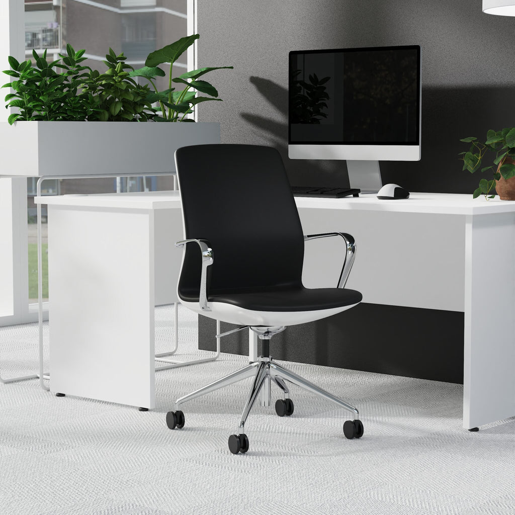 A modern black office chair with a white base and chrome armrests, positioned in front of a white desk with a computer monitor, plants, and a grey wall.