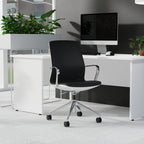 A modern black office chair with a white base and chrome armrests, positioned in front of a white desk with a computer monitor, plants, and a grey wall.