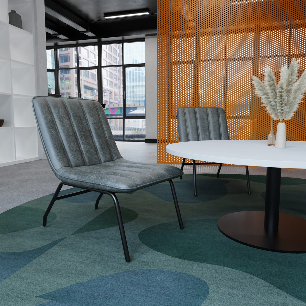 A modern grey upholstered lounge chair with black metal legs, placed in a contemporary office or lounge setting with a round white table, a second matching chair, and a patterned blue-green rug.