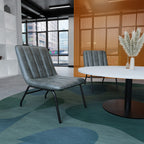 A modern grey upholstered lounge chair with black metal legs, placed in a contemporary office or lounge setting with a round white table, a second matching chair, and a patterned blue-green rug.
