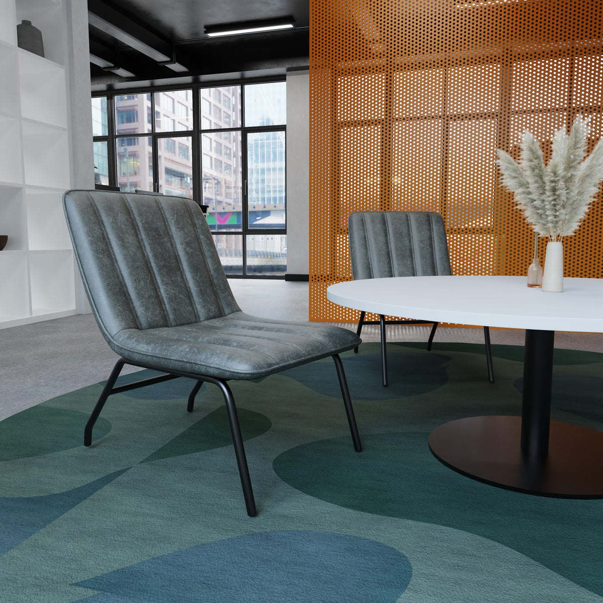 A modern grey upholstered lounge chair with black metal legs, placed in a contemporary office or lounge setting with a round white table, a second matching chair, and a patterned blue-green rug.
