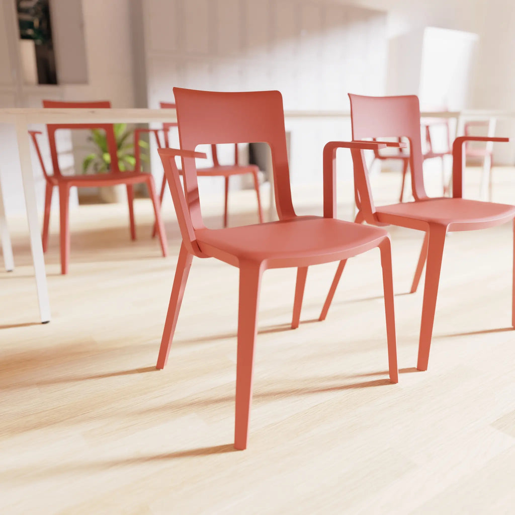 A modern terracotta red plastic dining chair with armrests, shown in a bright, sunlit room with light wood floors and other matching chairs in the background.