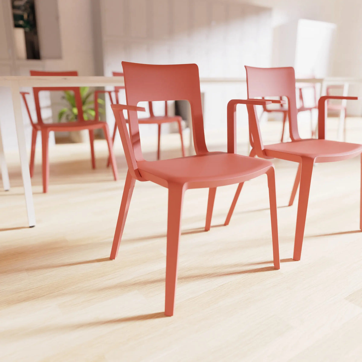 A modern terracotta red plastic dining chair with armrests, shown in a bright, sunlit room with light wood floors and other matching chairs in the background.