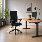 A modern home office setup featuring a black ergonomic office chair next to a wooden height-adjustable desk with a computer monitor, desk lamp, and small potted plant, set against a neutral beige wall and light wood flooring.