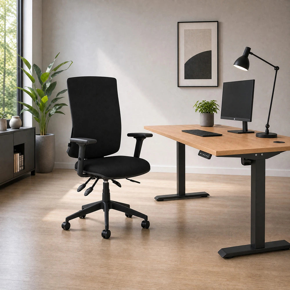 A modern home office setup featuring a black ergonomic office chair next to a wooden height-adjustable desk with a computer monitor, desk lamp, and small potted plant, set against a neutral beige wall and light wood flooring.