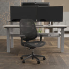 A modern black mesh office chair with a five-star wheeled base, positioned in front of a light grey standing desk with dual computer monitors in a home office setting.