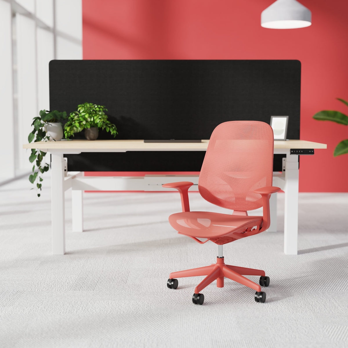 A modern coral pink ergonomic office chair with a mesh back and five-wheel base, positioned in front of a desk with a black screen in a bright office setting.
