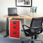 The desk styled in an office setting with a black mesh office chair, grey filing cabinet with red doors, a laptop, and a mint green magazine rack.
