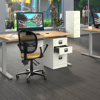 A modern office setup featuring a white three-drawer pedestal file cabinet with an open drawer, paired with a black mesh office chair and a light wood desk surface. The scene is set in a bright office with grey flooring, other desks, and blurred people visible in the background.