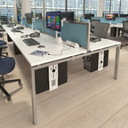 A modern office desk setup with a white desktop, silver metal frame, light blue privacy dividers, and black under-desk storage cabinets, shown in a bright office environment with office chairs and computer monitors.