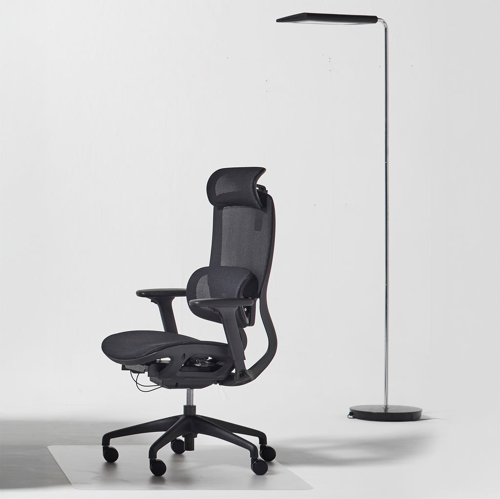 A modern black ergonomic office chair with a mesh back and headrest, positioned next to a slim black floor lamp on a light gray studio background.