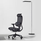 A modern black ergonomic office chair with a mesh back and headrest, positioned next to a slim black floor lamp on a light gray studio background.