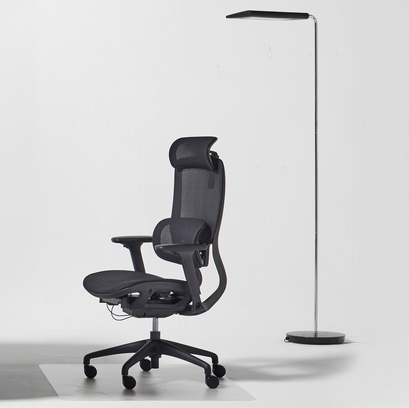 A modern black ergonomic office chair with a mesh back and headrest, positioned next to a slim black floor lamp on a light gray studio background.