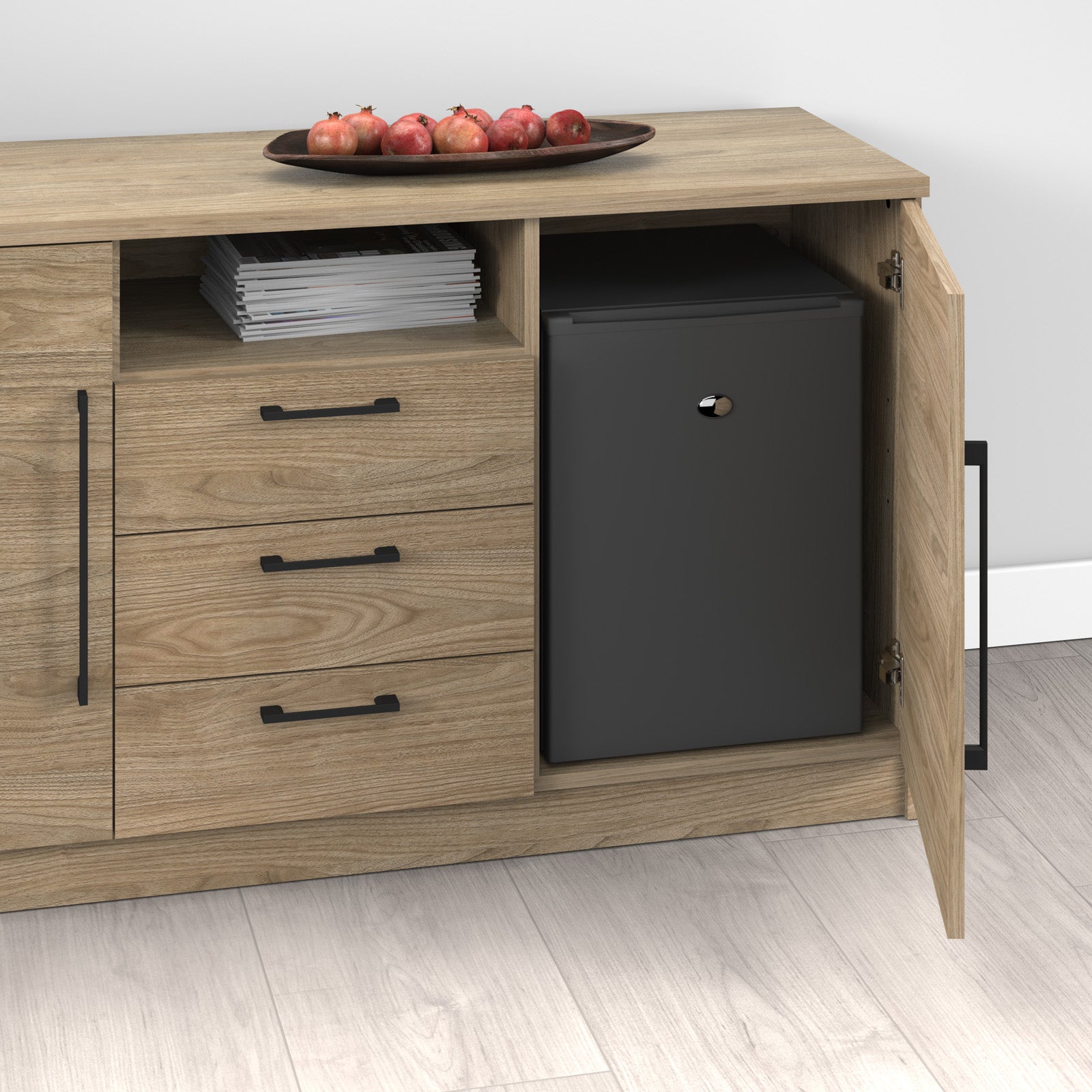 A light wood storage cabinet with three drawers, open shelves, and a door pulled open to reveal a small black refrigerator inside. A tray of red apples and a stack of magazines sit on top.