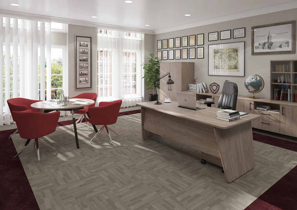 A modern office interior featuring a large light wood desk with a black office chair, a round dining table with four red chairs, and a herringbone-patterned grey rug. The room is styled with framed wall art, a bookshelf, and a potted plant.