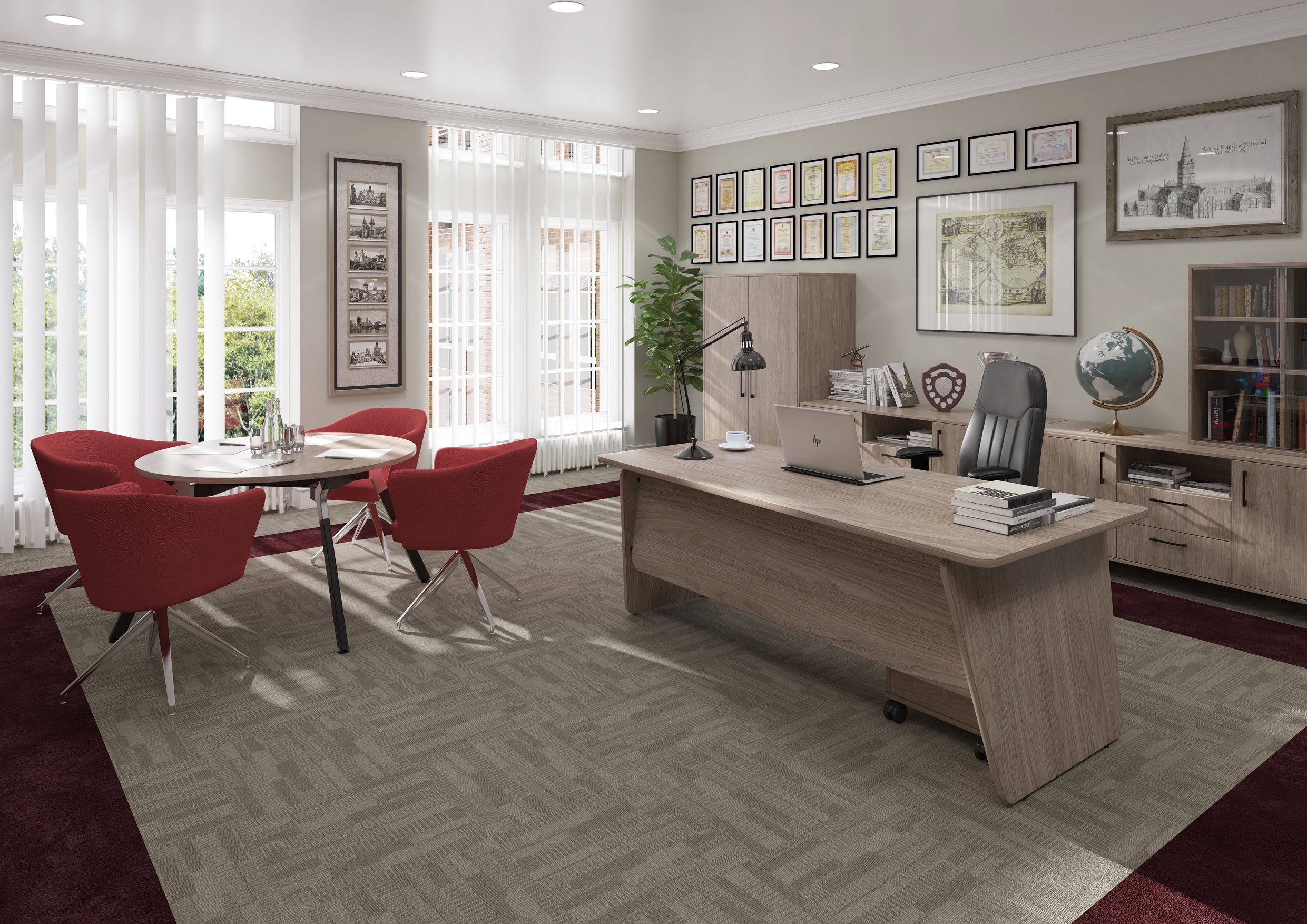 A modern office interior featuring a large light wood desk with a black office chair, a round dining table with four red chairs, and a herringbone-patterned grey rug. The room is styled with framed wall art, a bookshelf, and a potted plant.