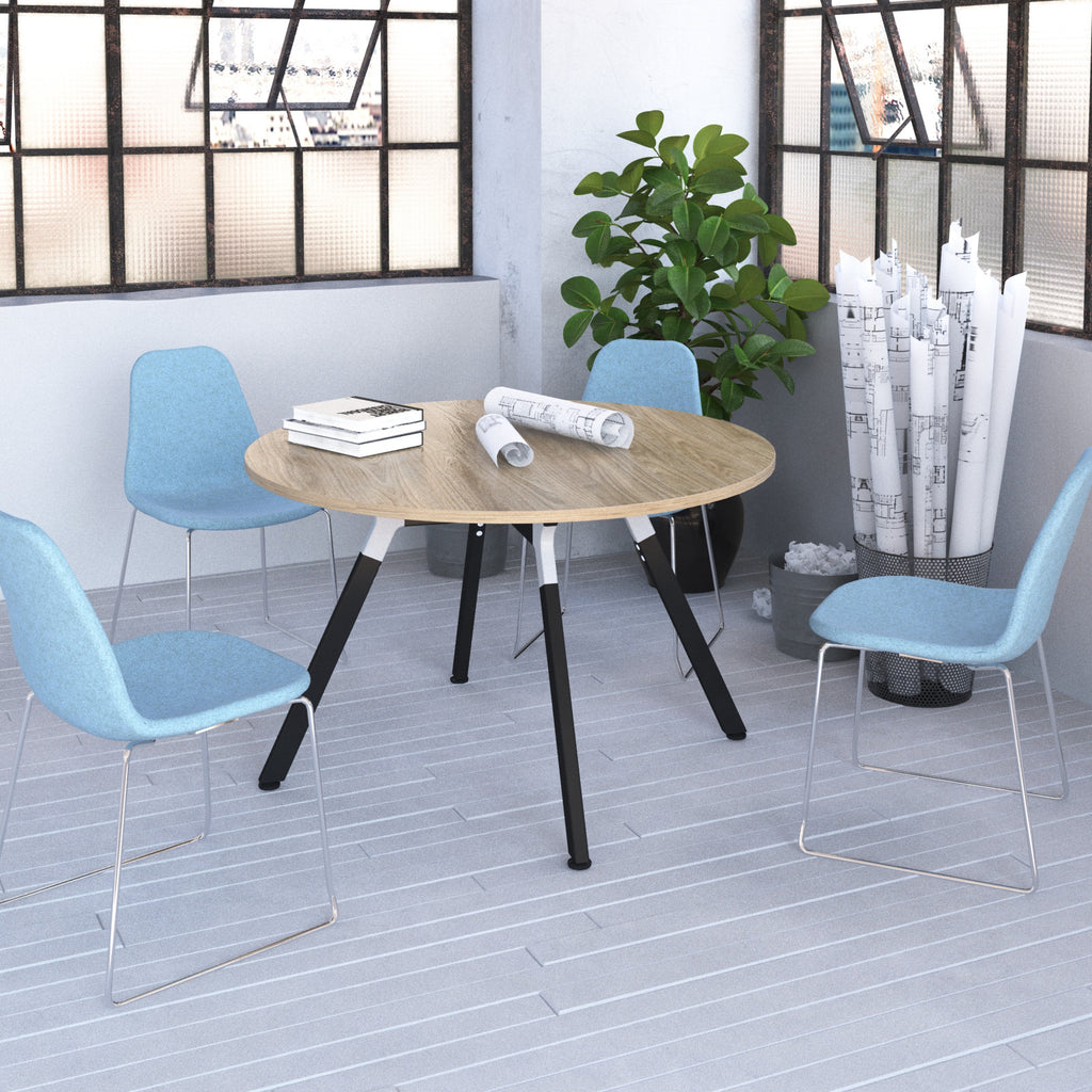 A round wooden table with black angled legs is surrounded by four light blue chairs in a bright office setting with large black-framed windows, a potted plant, and rolled blueprints.