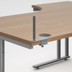 A close-up of a modern desk showing a light wood grain desktop with a curved cut-out, a grey metal support post, a cable management grommet, and a grey metal leg frame on a light grey background.