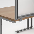 A close-up of a modern desk with a light wood grain tabletop and a grey metal frame supporting a clear glass panel.