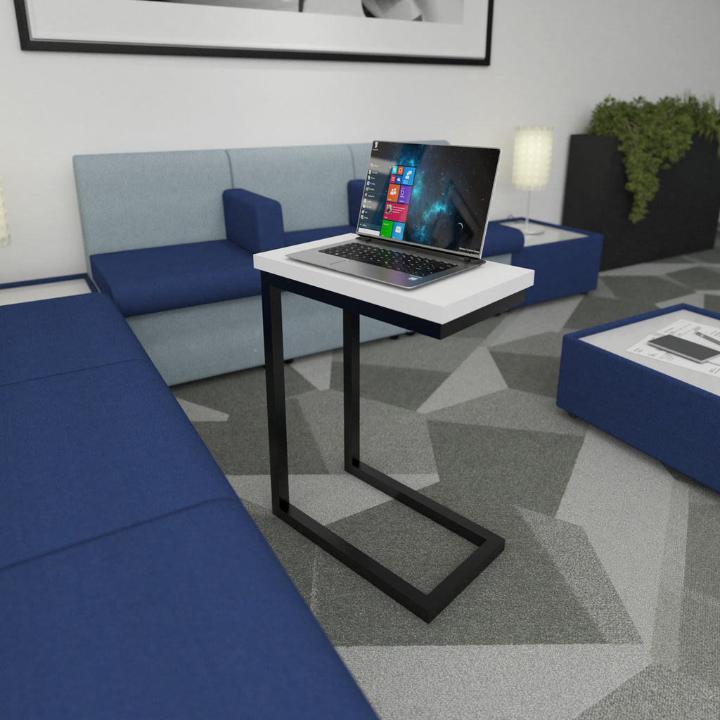 A modern standing laptop table with a white rectangular top and black metal frame, holding an open laptop in a contemporary lounge setting with blue seating and a patterned grey rug.