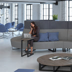 A woman sits on a curved grey modular office sofa with blue pillows, using a laptop on a small black side table. The scene is set in a modern office with blue chairs, large windows, and a round wooden coffee table in the foreground.
