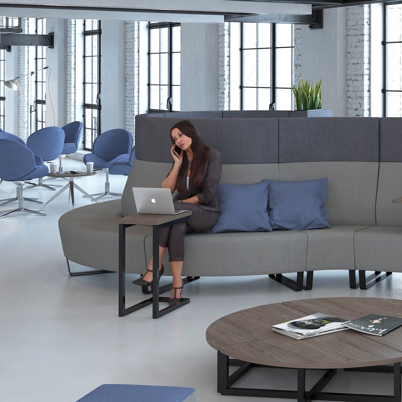 A woman sits on a curved grey modular office sofa with blue pillows, using a laptop on a small black side table. The scene is set in a modern office with blue chairs, large windows, and a round wooden coffee table in the foreground.