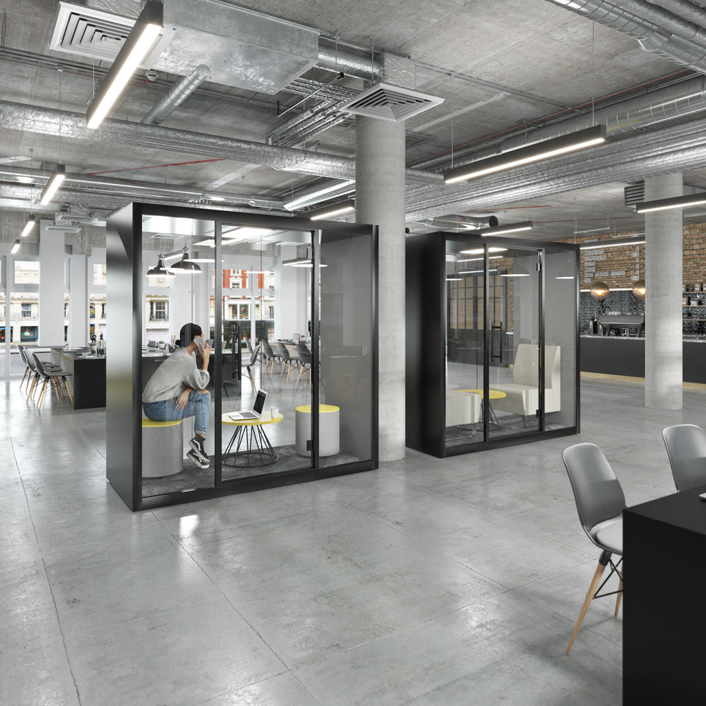 Modern office interior with two black glass privacy callout booths. One booth is occupied by a person sitting on a gray chair, while the other stands empty.