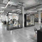 Modern office interior with two black glass privacy callout booths. One booth is occupied by a person sitting on a gray chair, while the other stands empty.