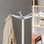 White coat rack with a beige coat against a gray wall, with a bookshelf and chair in the background.
