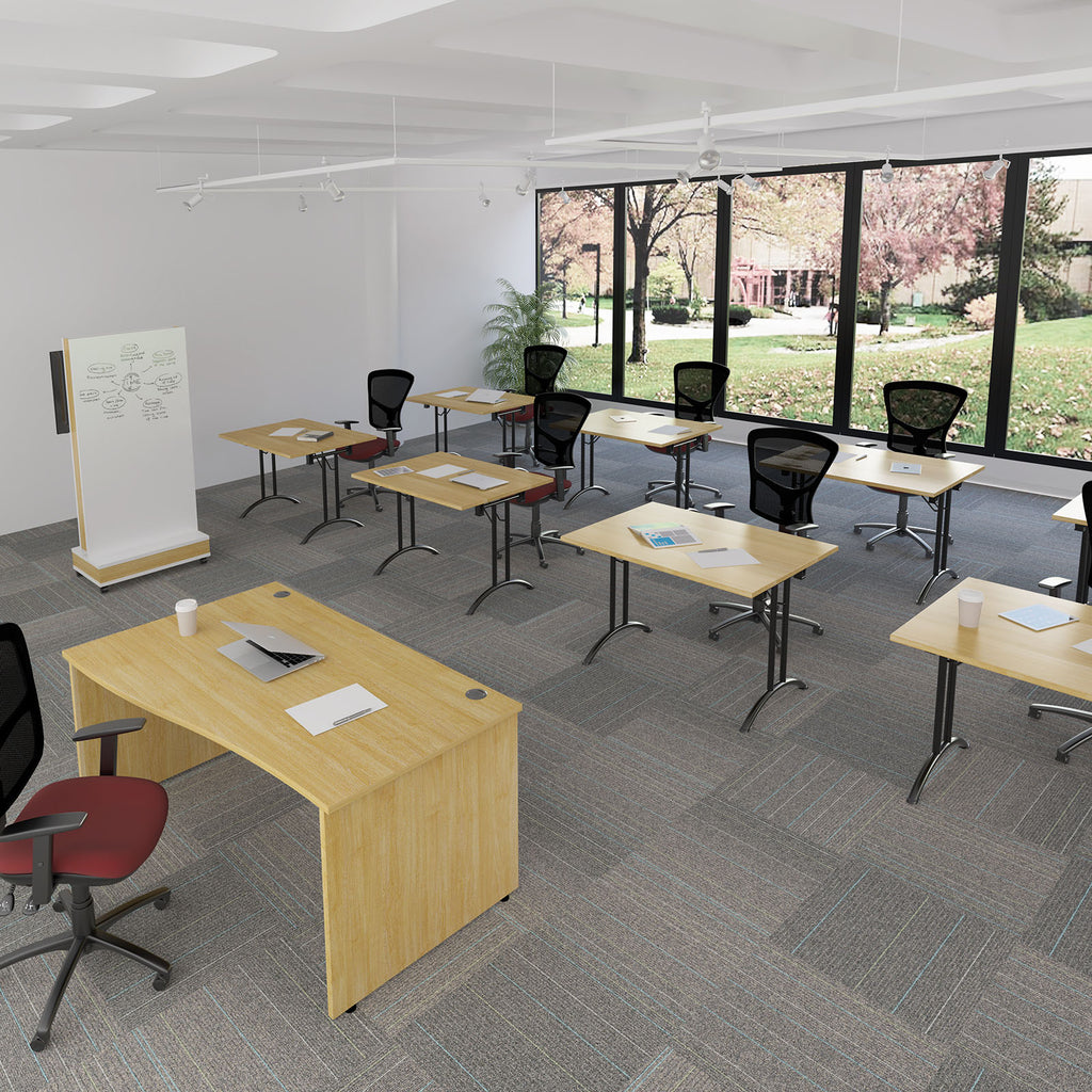 A modern classroom or office space with multiple light wood desks, black mesh office chairs, a whiteboard, and large windows overlooking a landscaped outdoor area.