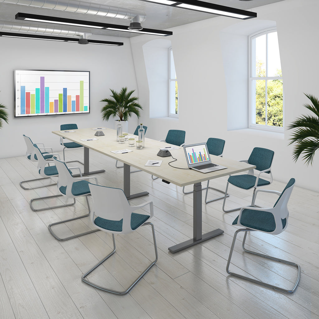 A modern conference room setup with a light wood rectangular meeting table surrounded by white and teal chairs with chrome metal legs. The room has white walls, large windows, ceiling lights, potted plants, and a wall-mounted screen showing a colorful bar chart.