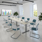A modern conference room setup with a light wood rectangular meeting table surrounded by white and teal chairs with chrome metal legs. The room has white walls, large windows, ceiling lights, potted plants, and a wall-mounted screen showing a colorful bar chart.