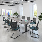 A modern conference room setup with a long rectangular desk and multiple mesh office chairs featuring dark grey frames and teal-blue seat cushions, placed in a bright white room with large windows and a framed multicolour bar chart on the wall.